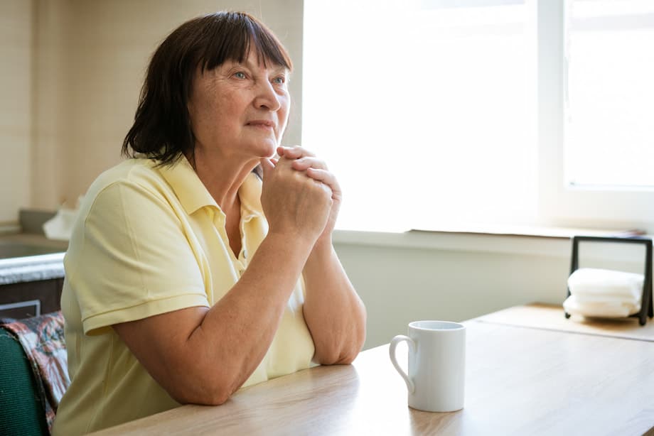 Elderly woman sitting at a table, hands clasped.